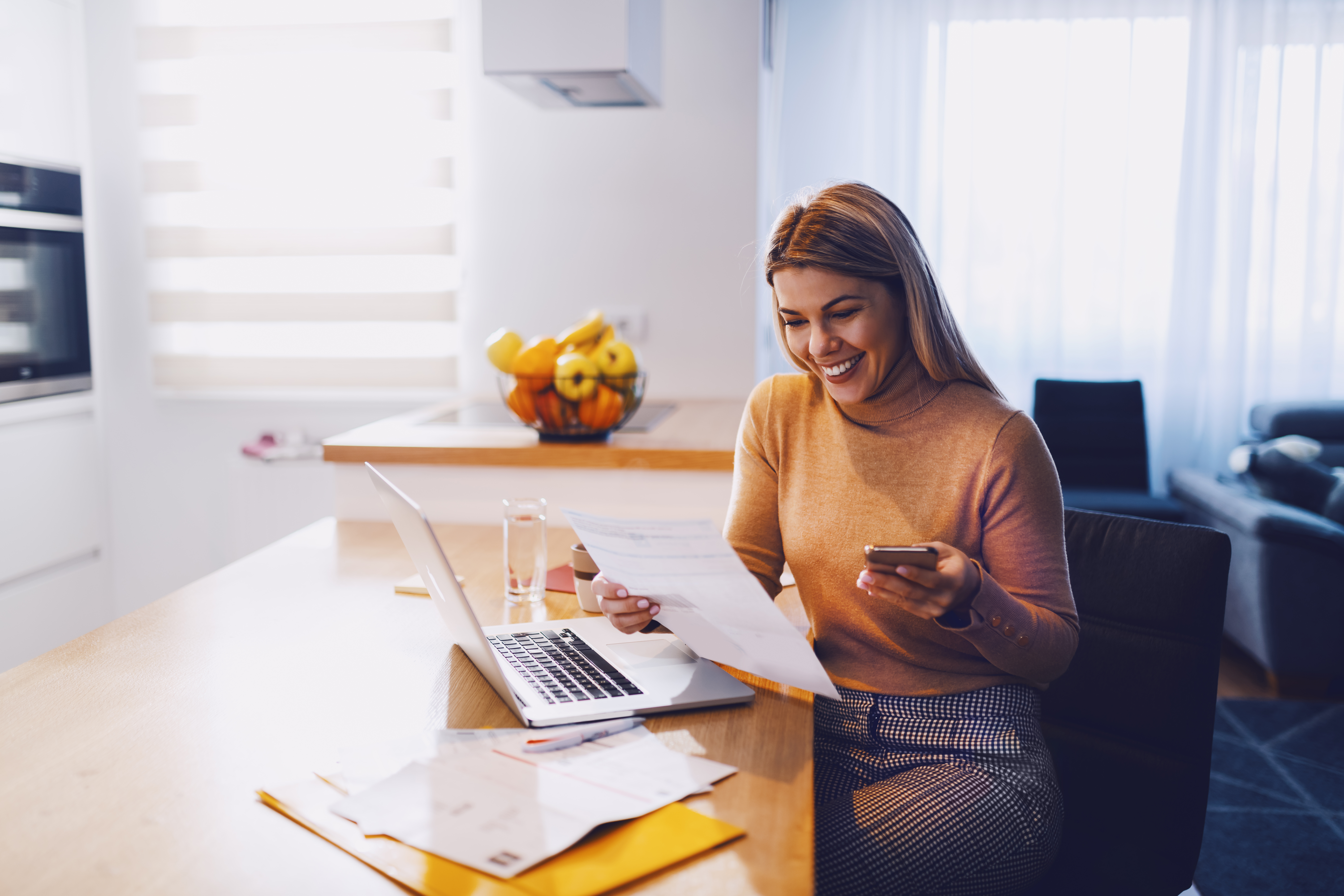 woman in sweater holding bills in one hand and in other smart phone. On table are laptop and bills. Apartment interior.