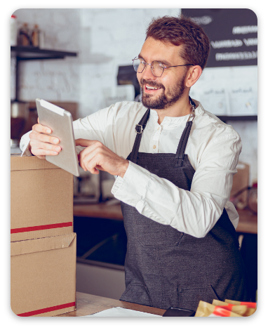 Joyful bearded man in apron using modern gadget and smiling while standing behind counter with delivery boxes
